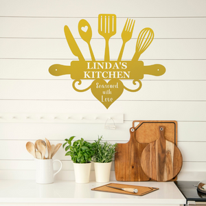 A personalized kitchen wall decal in a warm mustard yellow color, featuring kitchen utensils and the text 'LINDA'S KITCHEN' and 'Seasoned with Love'. The decal is displayed on a white shiplap wall above a kitchen counter with wooden cutting boards, potted green plants, and a white mug containing wooden spoons.