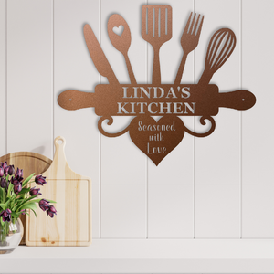 A rustic metal kitchen wall sign reading 'LINDA'S KITCHEN' and 'Seasoned with Love,' styled on a white paneled wall above a countertop with wooden boards and purple flowers.