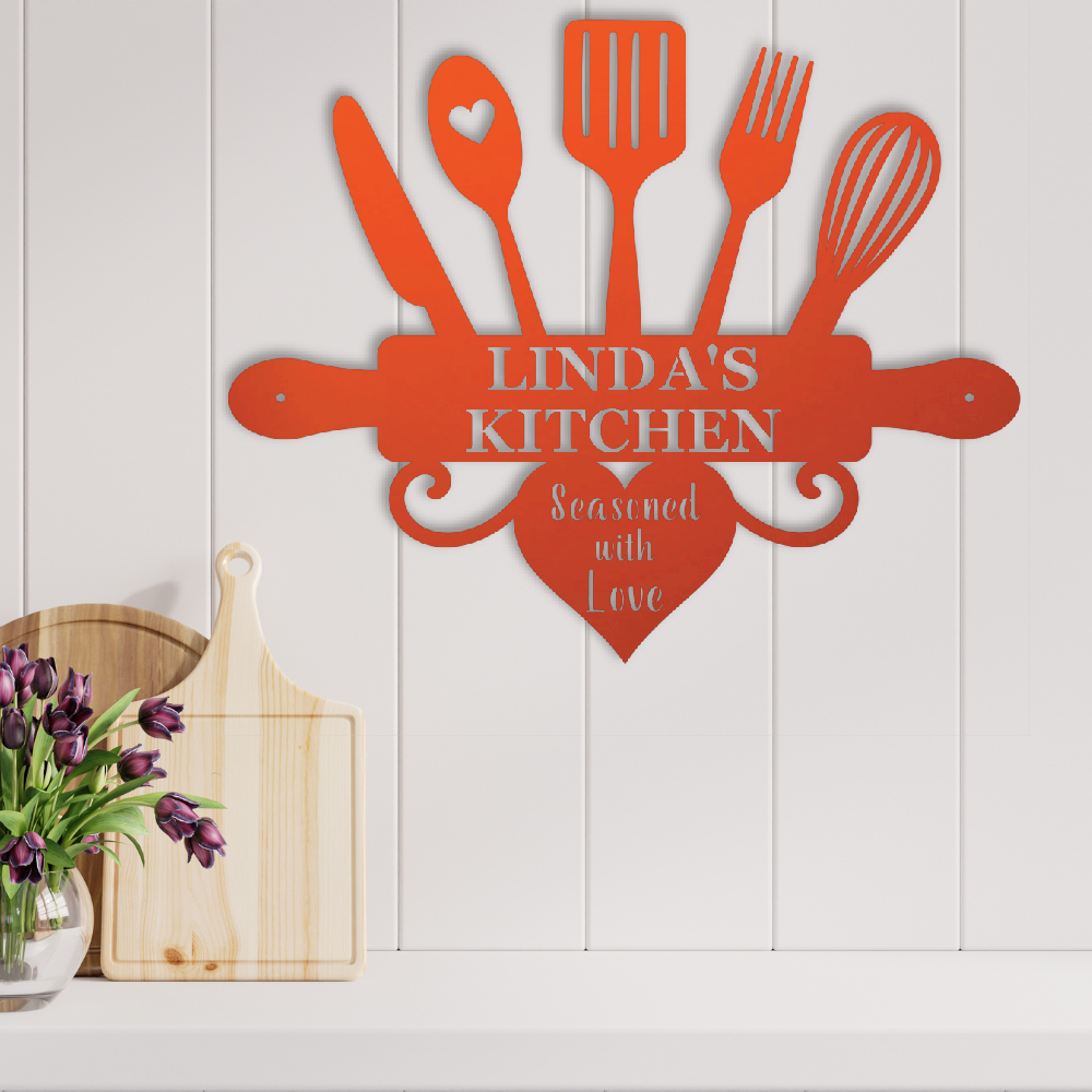 A personalized metal kitchen wall sign in a burnt orange-red color, featuring kitchen utensils and a rolling pin with the text 'LINDA'S KITCHEN' and 'Seasoned with Love'. The sign is mounted on a white paneled wall above a kitchen counter with wooden cutting boards and a vase of purple tulips.