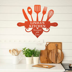 A red wall decal shaped like a kitchen utensil cluster with the text 'LINDA'S KITCHEN' and 'Seasoned with Love' displayed on a white shiplap wall above a kitchen counter with cutting boards, plants, and wooden spoons.