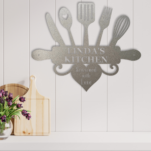 A silver glitter metal kitchen wall sign shaped like utensils with a heart and the words 'LINDA'S KITCHEN' and 'Seasoned with Love,' displayed on a white paneled wall above a countertop with wooden boards and flowers.