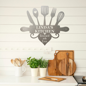 A silver metal kitchen wall sign reading 'LINDA'S KITCHEN' and 'Seasoned with Love' is displayed on a white shiplap wall above a kitchen counter with wooden cutting boards, potted green plants, and a white mug holding wooden utensils.