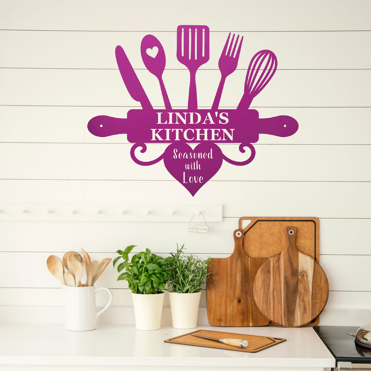 A purple kitchen-themed wall decal reading 'LINDA'S KITCHEN' and 'Seasoned with Love' is displayed on a white shiplap wall above a kitchen counter with wooden cutting boards, potted herbs, and utensils.