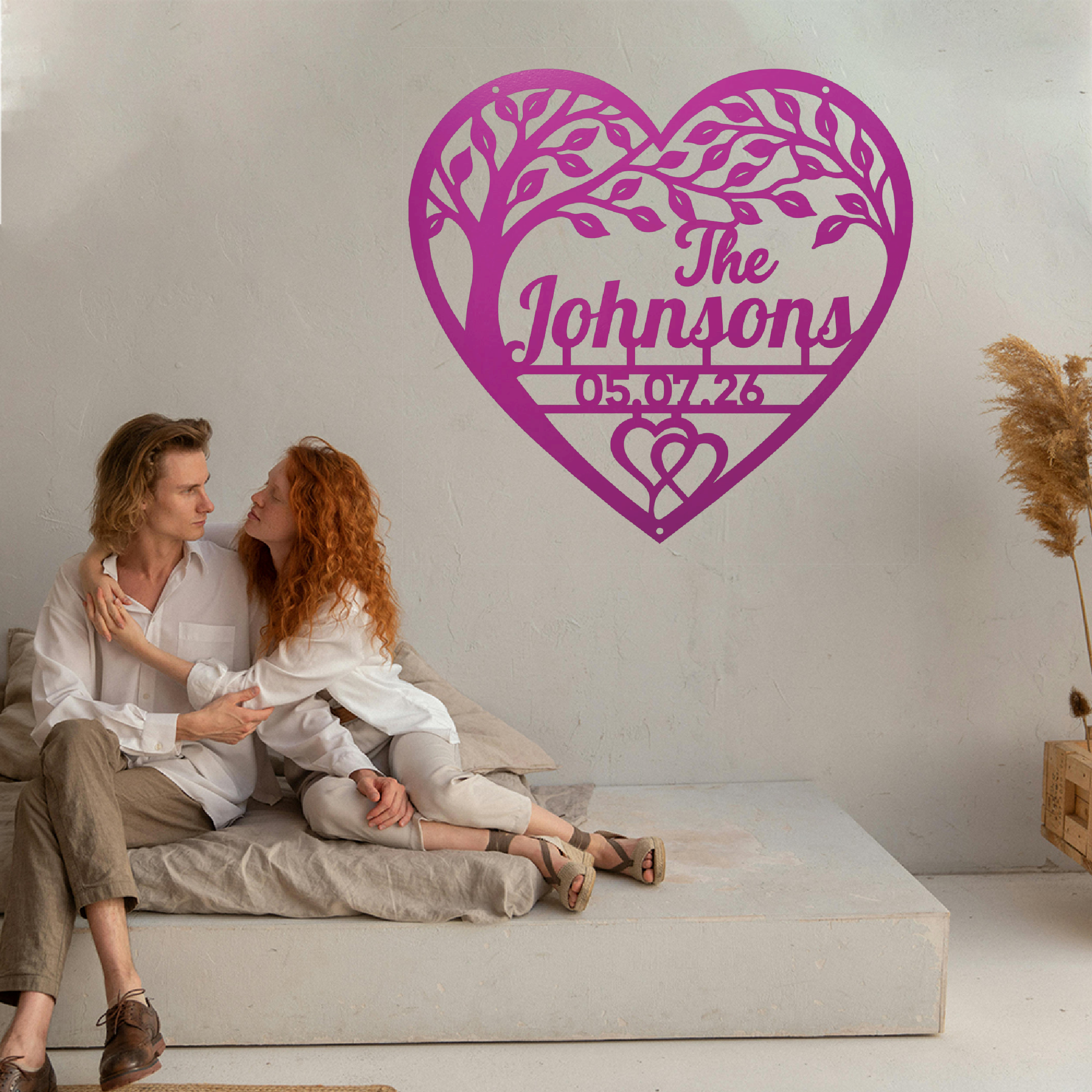 A couple sits on a low concrete bench against a light wall, with a large magenta heart-shaped tree silhouette and custom text reading 'The Johnsons' and '05.07.