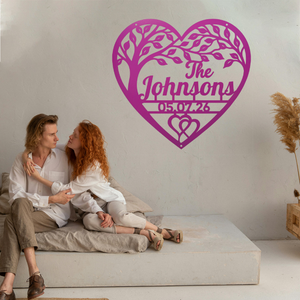 A couple sits on a low concrete bench against a light wall, with a large magenta heart-shaped tree silhouette and custom text reading 'The Johnsons' and '05.07.