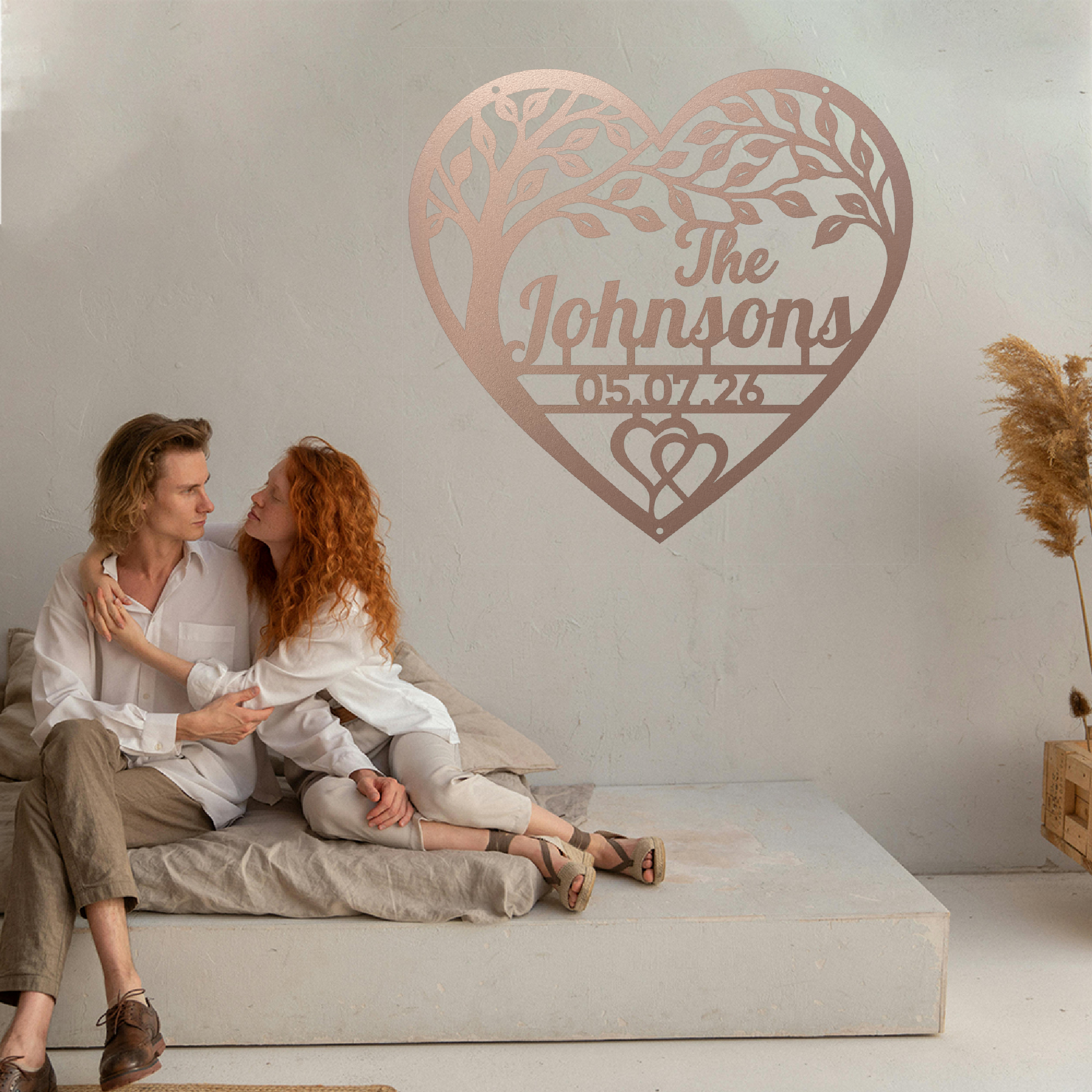 A couple sits closely together on a low concrete bench in a minimalist room, with a large personalized rose-gold heart-shaped tree wall art piece above them featuring a custom name and date.